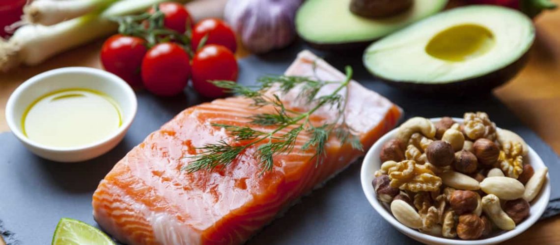 Table top still life of foods high in healthy fats such as Salmon, olive oil, nuts and avocados with vegetables and herbs.