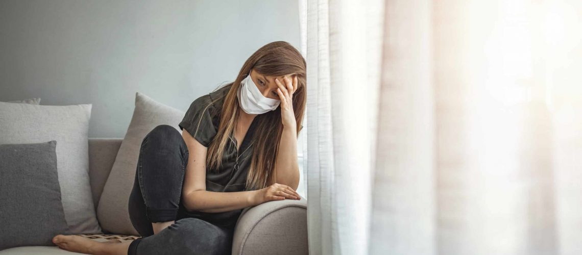 Sad lonely girl isolated stay at home in protective sterile medical mask on face looking at window, bored woman because of Chinese pandemic coronavirus virus covid-19. Quarantine, prevent infection