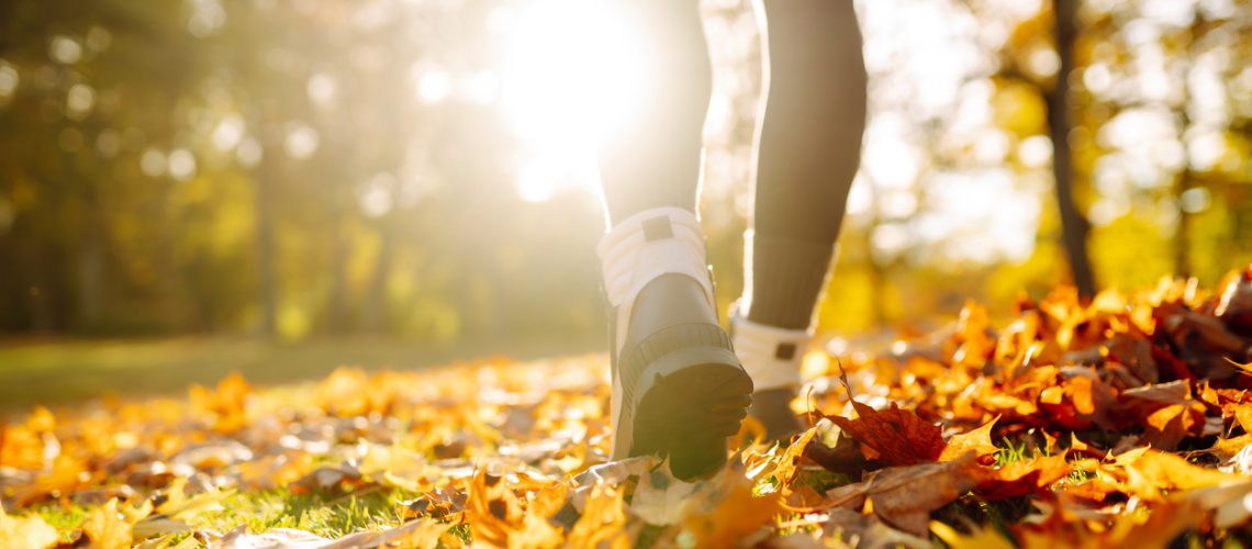 Close-up of female legs in hiking boots walks on ground with yellow-orange dry fall leaves during autumn season in park or forest. Feet walking in outdoor nature. Healthy lifestyle on leisure activity