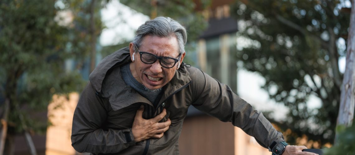 Senior man suddenly feeling a sharp pain in his chest while running, causing him to slow down and griping a nearby pole for support. With a trembling hand, he quickly glances down at his smartwatch, hoping to check his heart rate and assess his condition