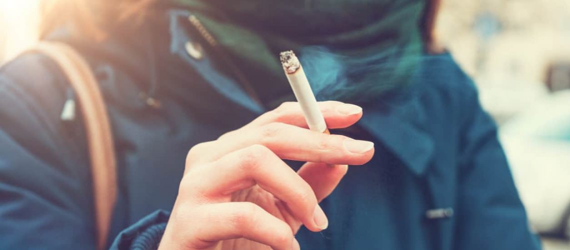 Young woman enjoying a cigarette outdoors holding it between her fingers, low angle view against the chest of a warm autumn jacket in a smoking and tobacco concept